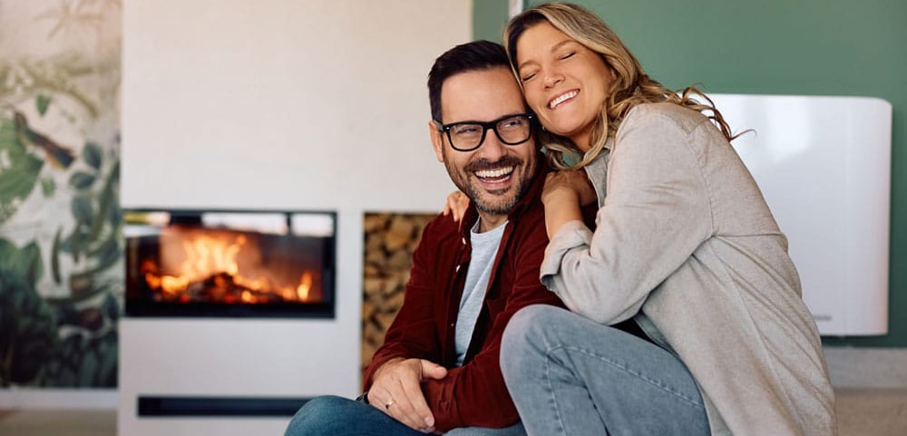 Furnace Repair Homeowning couple hug and smile with fireplace on in the background of their living room.
